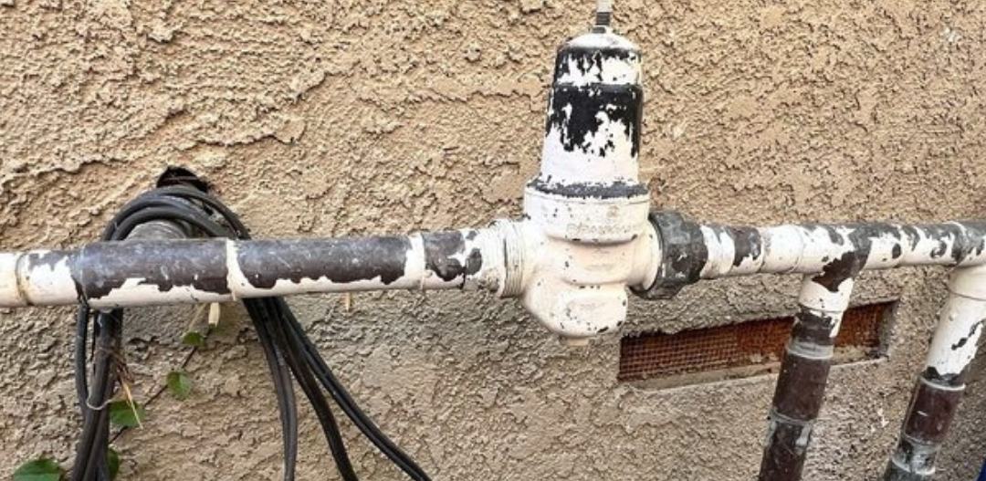 Aging threaded steel water supply pipe and pressure regulator with peeling paint on the outside wall of a Southern California home